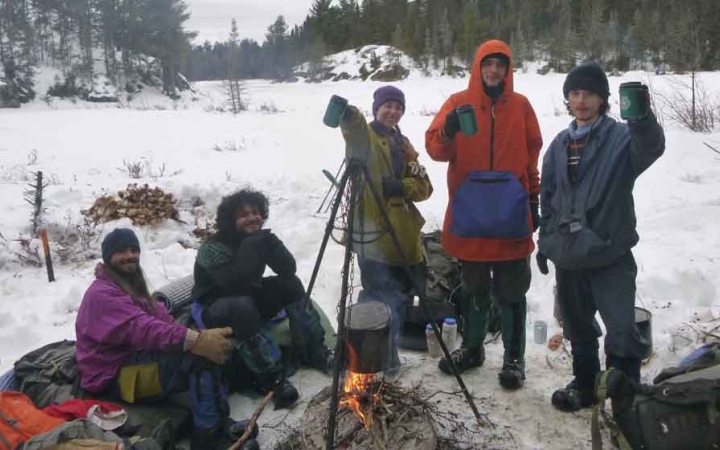 a group of gap year students gathered around a campfire in a snowy landscape raise thermoses in the air